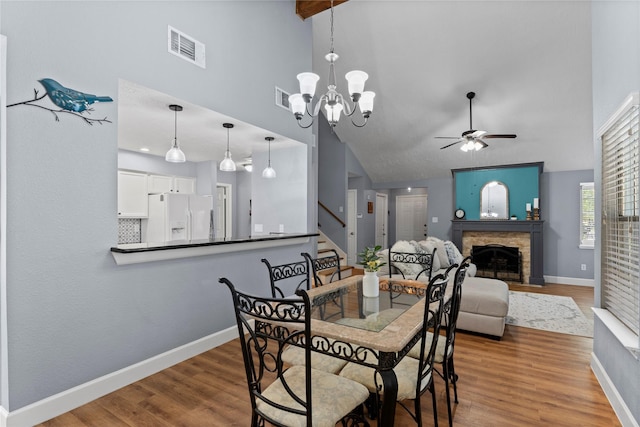 dining area featuring a stone fireplace, hardwood / wood-style floors, high vaulted ceiling, and ceiling fan with notable chandelier