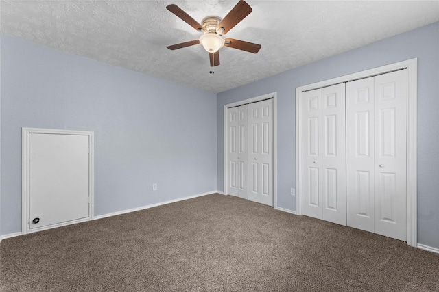 unfurnished bedroom with a textured ceiling, ceiling fan, dark carpet, and two closets