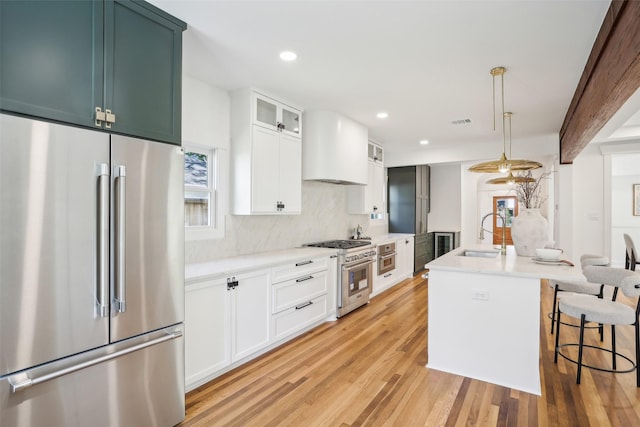 kitchen featuring appliances with stainless steel finishes, premium range hood, sink, a center island with sink, and light hardwood / wood-style floors