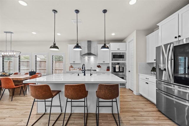 kitchen with an island with sink, stainless steel appliances, decorative light fixtures, wall chimney range hood, and white cabinets