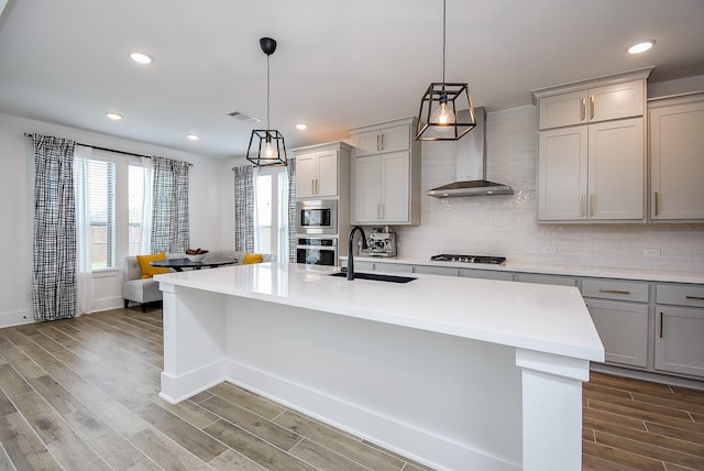 kitchen with wall chimney exhaust hood, gray cabinets, decorative backsplash, an island with sink, and stainless steel appliances