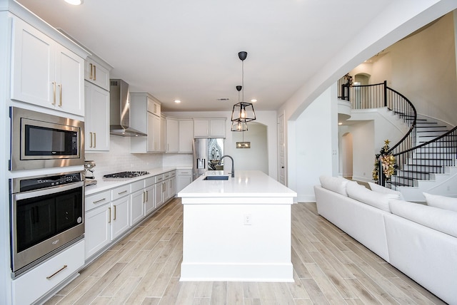 kitchen with sink, wall chimney exhaust hood, an island with sink, decorative light fixtures, and appliances with stainless steel finishes