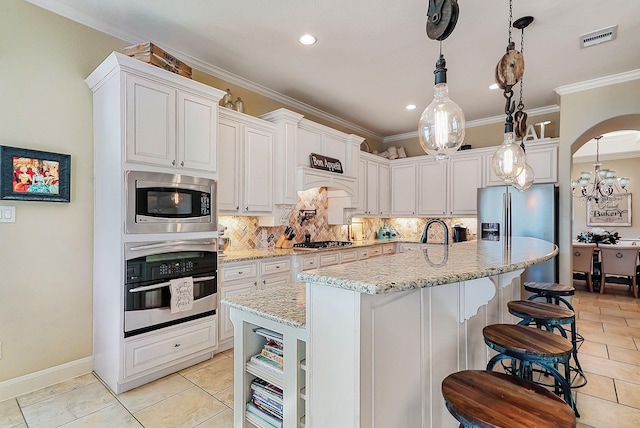 kitchen with light stone counters, stainless steel appliances, a kitchen island with sink, white cabinetry, and hanging light fixtures