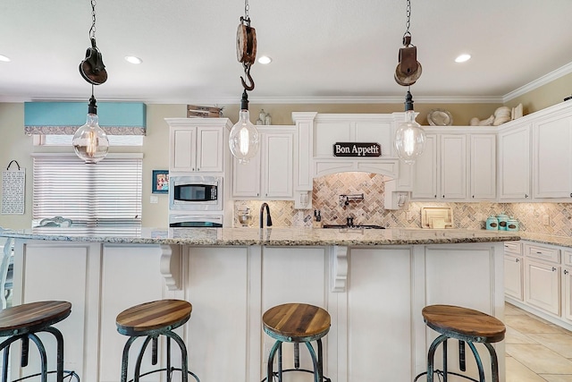 kitchen featuring light stone counters, ornamental molding, a kitchen island with sink, decorative light fixtures, and white cabinets