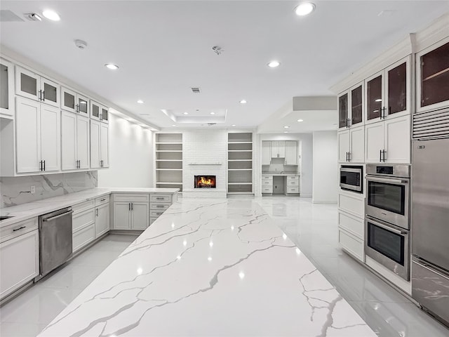 kitchen with light stone counters, built in shelves, a large fireplace, built in appliances, and white cabinetry