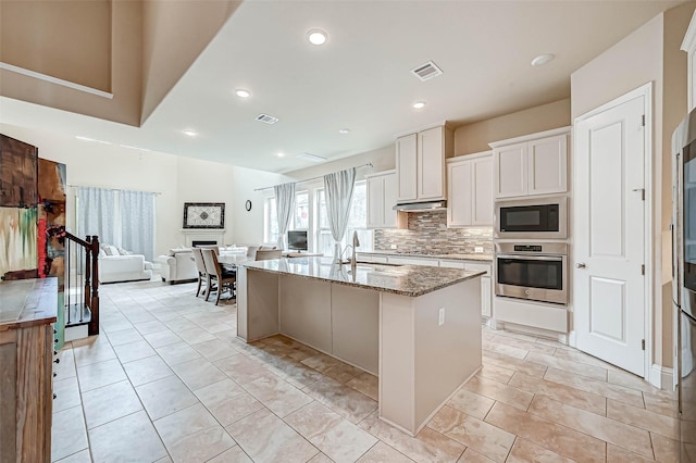 kitchen with built in microwave, stainless steel oven, light stone counters, an island with sink, and white cabinets