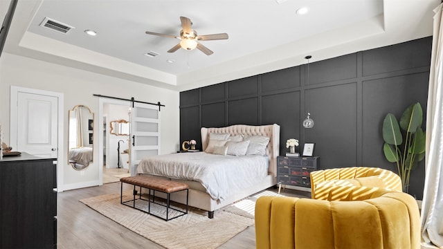 bedroom featuring a barn door, light hardwood / wood-style floors, a raised ceiling, and ceiling fan