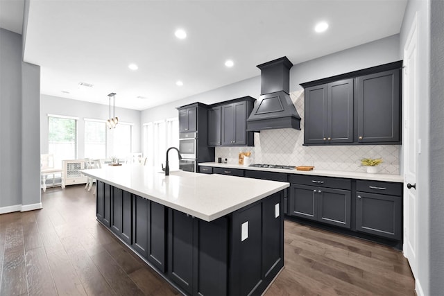 kitchen with decorative light fixtures, custom exhaust hood, stainless steel gas cooktop, dark wood-type flooring, and an island with sink