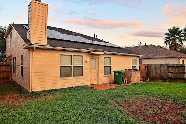 back of house at dusk with a fenced backyard, roof mounted solar panels, a chimney, and a yard