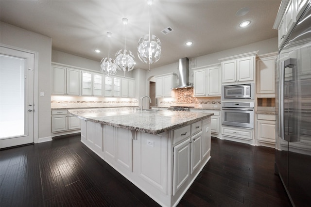 kitchen with a center island with sink, white cabinets, built in appliances, wall chimney exhaust hood, and dark hardwood / wood-style floors