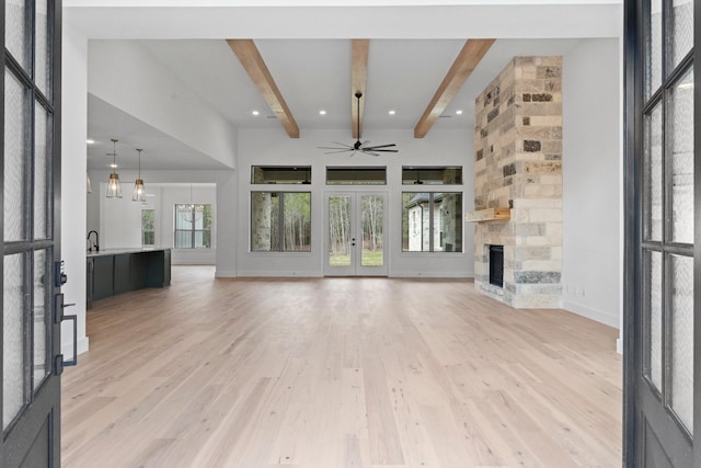 unfurnished living room with french doors, ceiling fan with notable chandelier, a fireplace, beamed ceiling, and light hardwood / wood-style floors