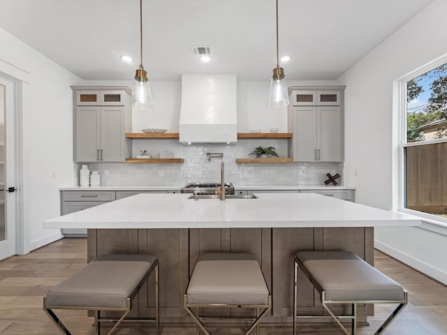 kitchen with decorative light fixtures, wood-type flooring, premium range hood, and a kitchen island with sink