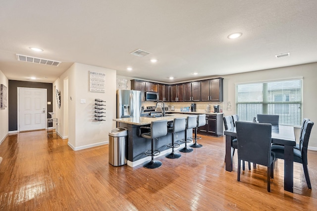 kitchen featuring stainless steel appliances, a kitchen breakfast bar, a kitchen island with sink, dark brown cabinets, and light wood-type flooring