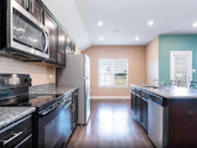 kitchen featuring light stone countertops, dark wood-type flooring, vaulted ceiling, dark brown cabinets, and appliances with stainless steel finishes