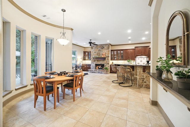 dining space featuring a stone fireplace, ceiling fan, crown molding, and light tile patterned flooring