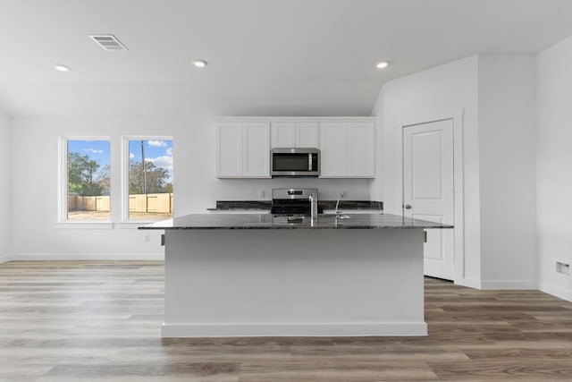 kitchen featuring appliances with stainless steel finishes, dark stone counters, a center island with sink, light hardwood / wood-style flooring, and white cabinets