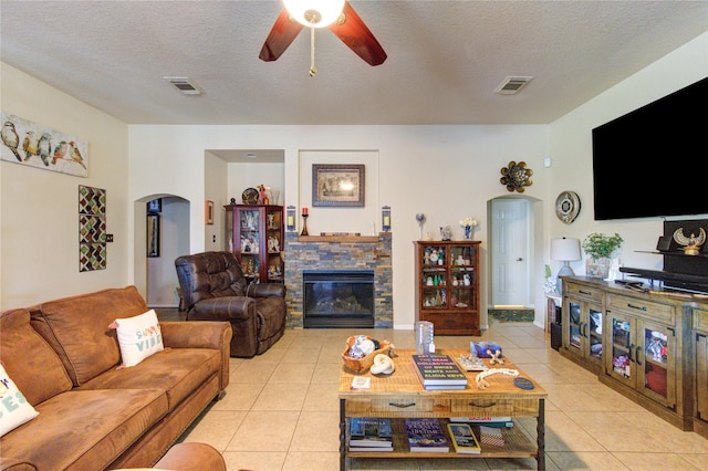 living room featuring ceiling fan, a stone fireplace, light tile patterned floors, and a textured ceiling