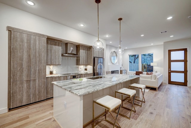 kitchen featuring pendant lighting, a center island with sink, wall chimney range hood, sink, and light hardwood / wood-style floors