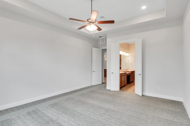 unfurnished bedroom featuring a raised ceiling, ensuite bath, ceiling fan, and light carpet