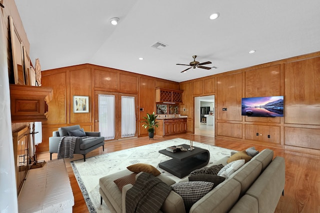 living room featuring light wood-type flooring, vaulted ceiling, ceiling fan, and wooden walls