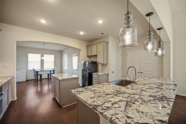 kitchen featuring sink, tasteful backsplash, pendant lighting, black refrigerator, and a center island with sink