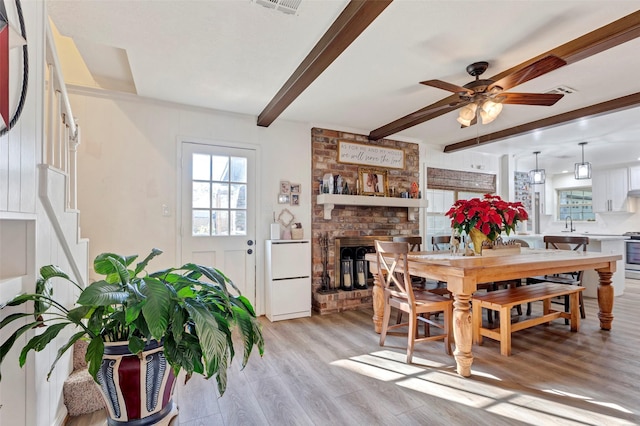 dining space with ceiling fan, beam ceiling, light wood-type flooring, and a brick fireplace