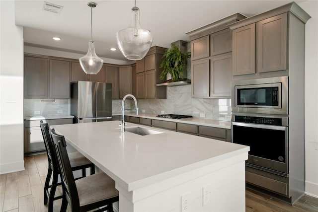 kitchen featuring sink, an island with sink, pendant lighting, stainless steel appliances, and wall chimney range hood