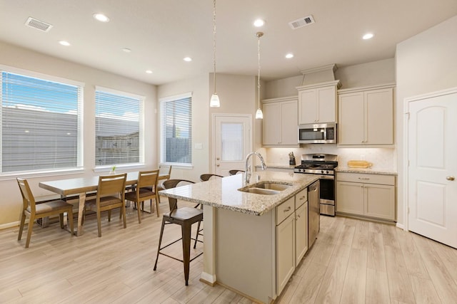 kitchen featuring a center island with sink, stainless steel appliances, hanging light fixtures, light stone counters, and sink