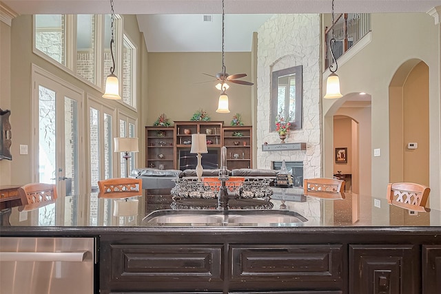 kitchen with ceiling fan, dishwasher, a stone fireplace, dark stone countertops, and dark brown cabinets