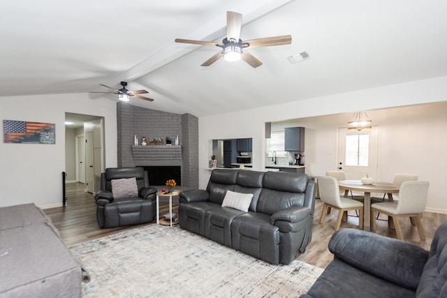 living room featuring ceiling fan with notable chandelier, a brick fireplace, vaulted ceiling with beams, and light hardwood / wood-style flooring