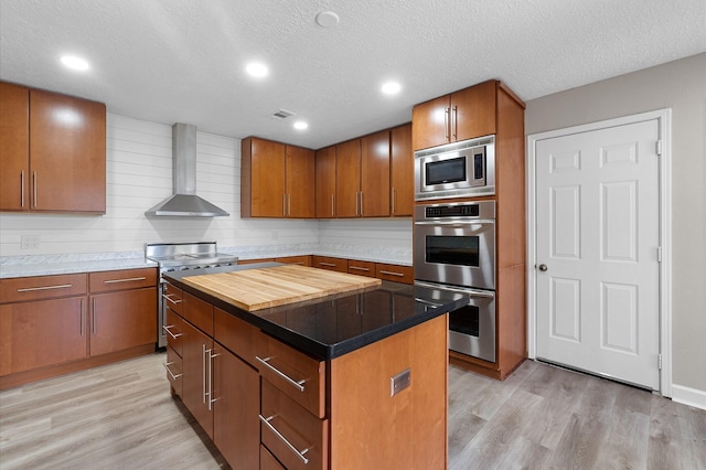 kitchen featuring light hardwood / wood-style flooring, wall chimney exhaust hood, a textured ceiling, appliances with stainless steel finishes, and a kitchen island