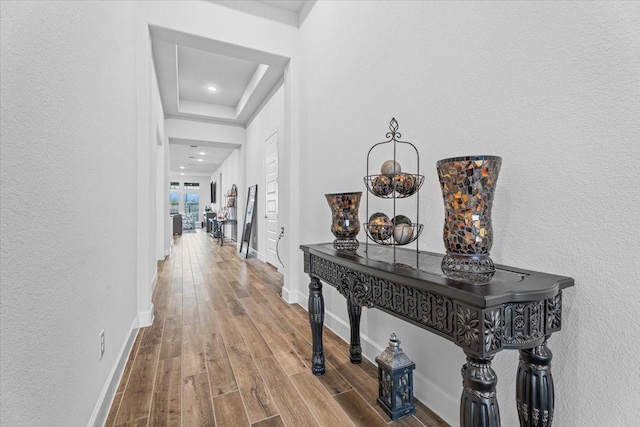 hallway featuring a textured wall, a raised ceiling, wood finished floors, and baseboards