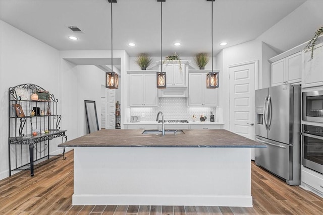 kitchen featuring hardwood / wood-style floors, appliances with stainless steel finishes, decorative light fixtures, white cabinetry, and a center island with sink