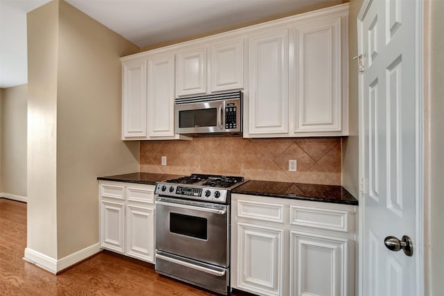 kitchen with appliances with stainless steel finishes, backsplash, dark stone counters, light hardwood / wood-style flooring, and white cabinets