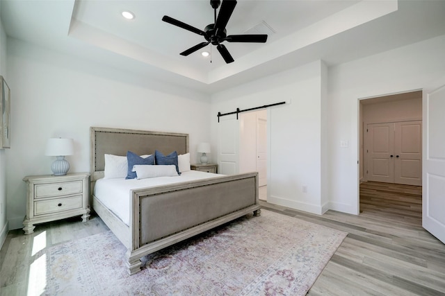 bedroom with light wood-type flooring, ceiling fan, a barn door, and a raised ceiling