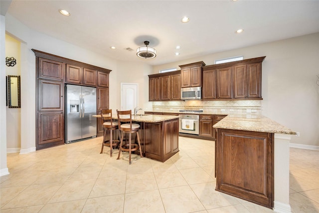 kitchen with stainless steel appliances, light stone counters, backsplash, a kitchen bar, and a kitchen island with sink