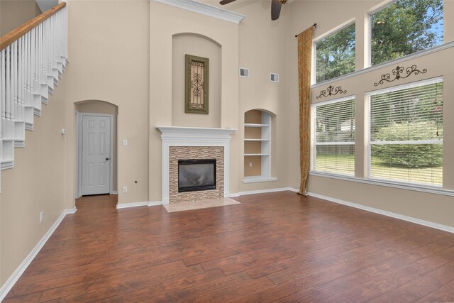 unfurnished living room with a high ceiling, built in shelves, ceiling fan, a fireplace, and dark hardwood / wood-style flooring