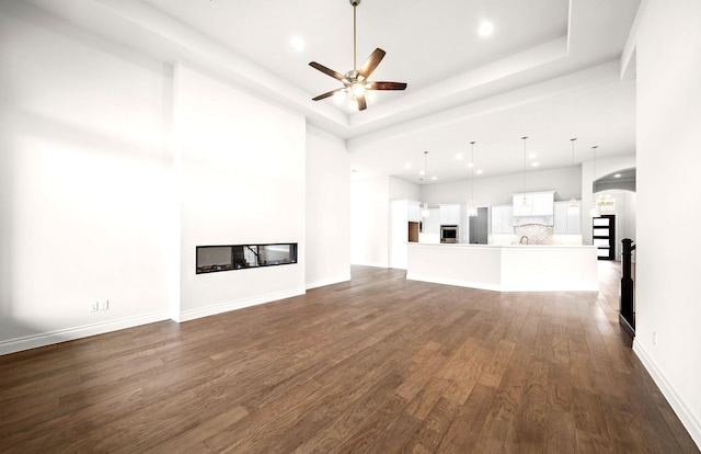 unfurnished living room featuring a glass covered fireplace, ceiling fan, a tray ceiling, and dark wood finished floors