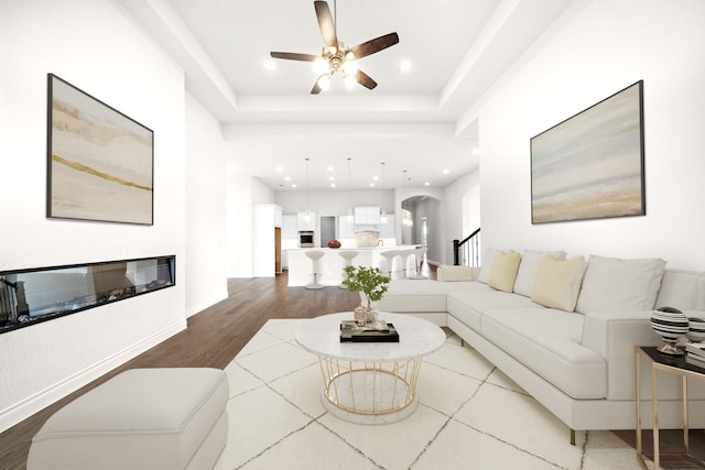 living area featuring a glass covered fireplace, wood finished floors, recessed lighting, a raised ceiling, and a ceiling fan