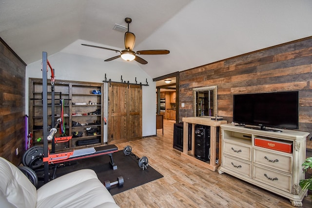 exercise area with ceiling fan, a barn door, wood walls, vaulted ceiling, and light wood-type flooring