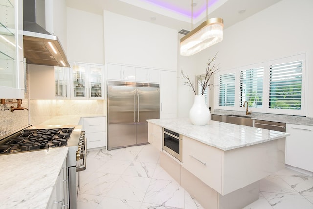 kitchen with high end appliances, wall chimney range hood, sink, light stone counters, and white cabinetry