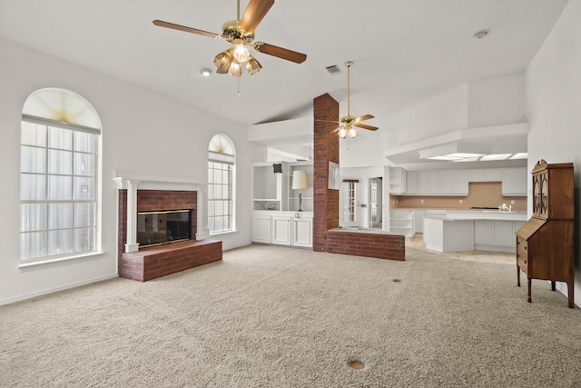 unfurnished living room with a brick fireplace, high vaulted ceiling, a ceiling fan, visible vents, and light colored carpet