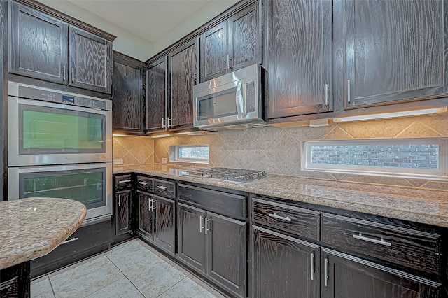 kitchen featuring light stone countertops, stainless steel appliances, tasteful backsplash, dark brown cabinets, and light tile patterned floors