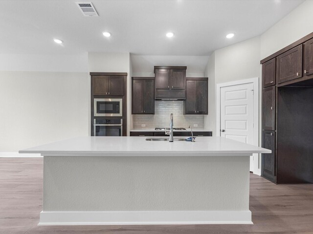 kitchen featuring stainless steel oven, a center island with sink, sink, built in microwave, and dark brown cabinetry