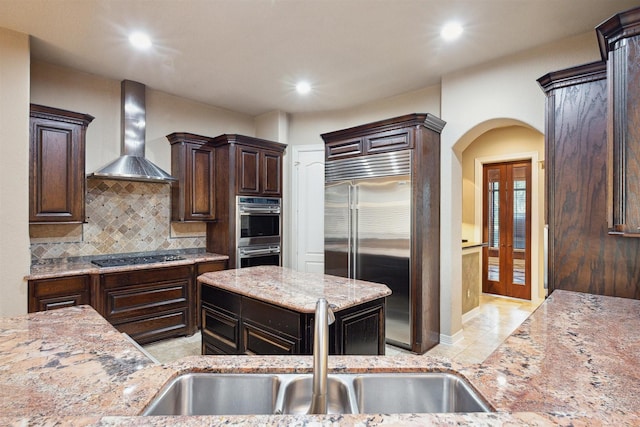 kitchen with dark brown cabinetry, light stone countertops, sink, wall chimney exhaust hood, and stainless steel appliances