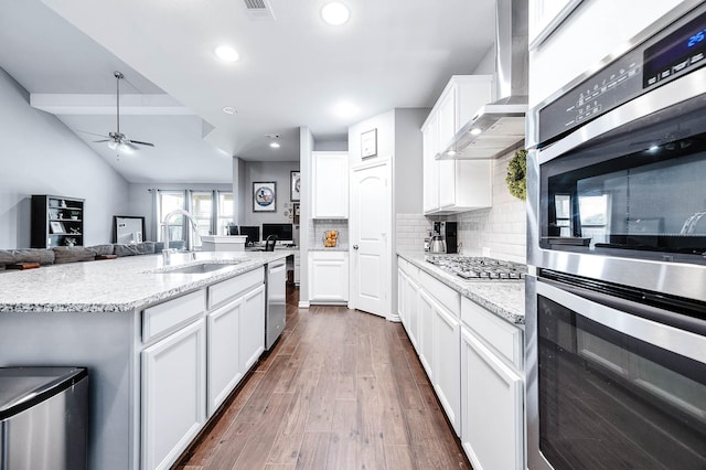 kitchen featuring white cabinetry, sink, stainless steel appliances, wall chimney range hood, and an island with sink