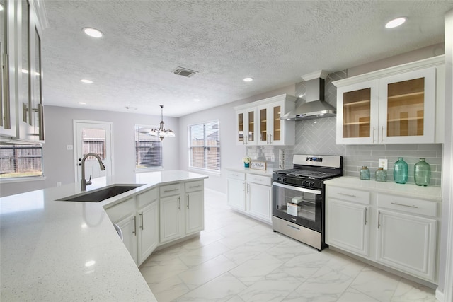 kitchen featuring white cabinetry, sink, hanging light fixtures, wall chimney range hood, and stainless steel gas range