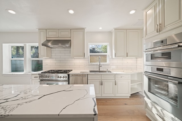 kitchen with wall chimney range hood, sink, light stone countertops, appliances with stainless steel finishes, and light hardwood / wood-style floors