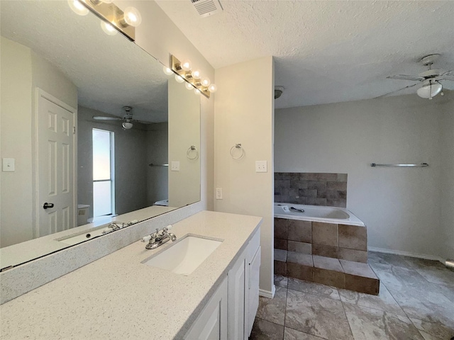 bathroom with vanity, a textured ceiling, ceiling fan, tiled bath, and toilet