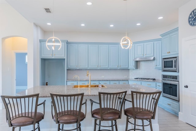 kitchen with appliances with stainless steel finishes, tasteful backsplash, a kitchen island with sink, sink, and hanging light fixtures